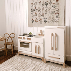 Milton & Goose Essential Play Kitchen in white along side the refrigerator in white. The items are set in a playroom with a child sized table and chairs.