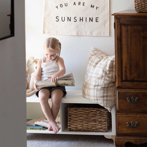 The wooden Archie Bench by Milton & Goose sits inside a child's room and features an oversized pillow and stuffies on top. A young girl sits on the bench reading a book.