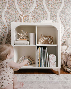 The wooden Archie Bookshelf by Milton & Goose is featured inside a nursery with baby toys and picture books stored inside. A young baby sits in front of the bookshelf reaching for some toys.