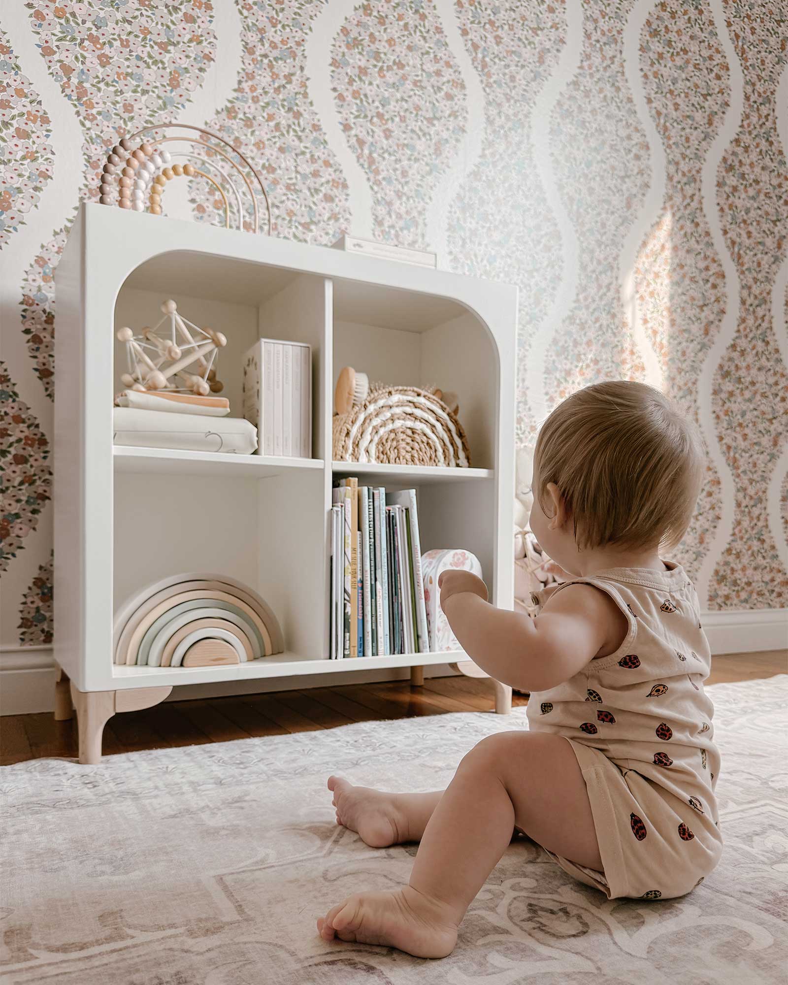 The wooden Archie Bookshelf by Milton & Goose is featured inside a nursery with baby toys and picture books stored inside. A young baby sits in front of the bookshelf reaching for some toys.