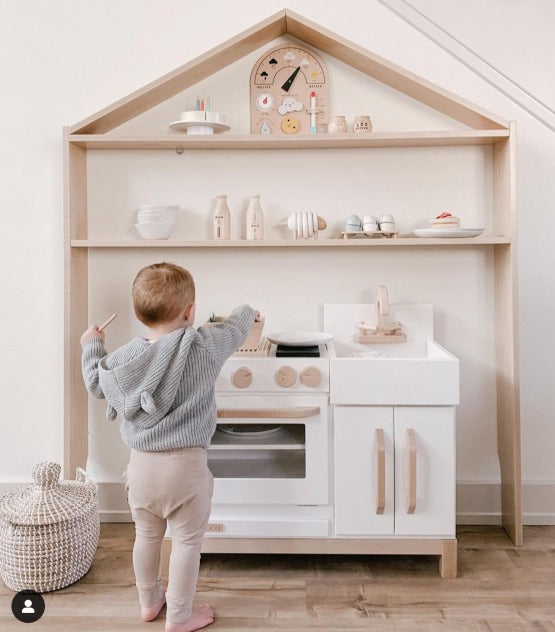 Child playing in front of Milton & Goose Essential Play kitchen in white. Kitchen set under the Milton & Goose House Shelf in Natural. Credit to IG profile @mandinelson_