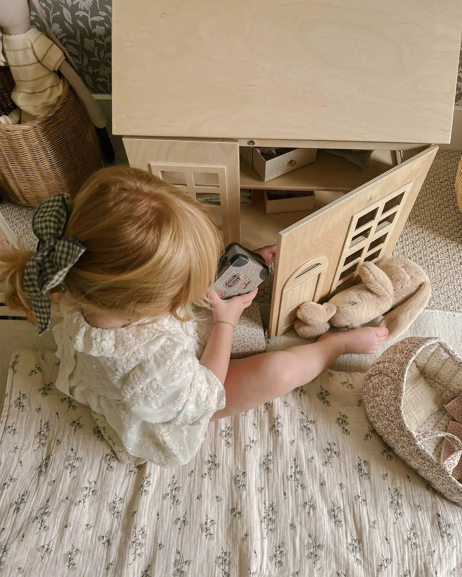 A child is shown playing with a natural wooden Hudson Dollhouse in a play room. 