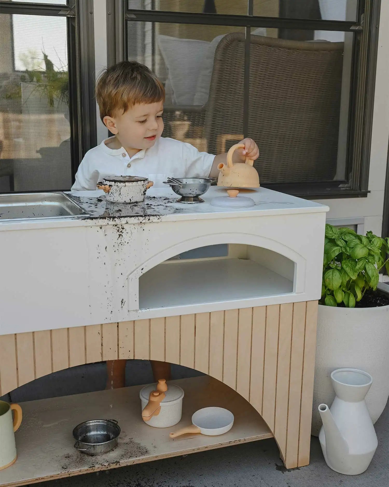 A child happily plays with the Kai Mud Kitchen while pouring mud and water into play pots and pans. 