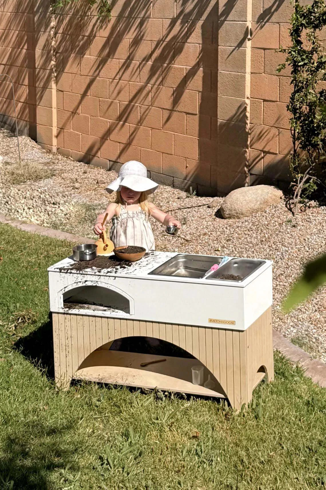 Girl plays at mud kitchen in yard. One sink basin is filled with water and the other has dirt. There is water and mud all over the countertop and dripping down the sides.