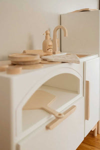 A close up look of the Milton & Goose wood Luca Play Kitchen in white. Image shows the pizza paddle sitting inside the pizza oven and various play food pieces sit on top of the kitchen.