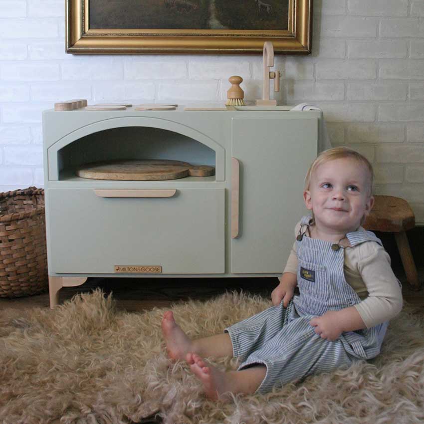 A baby smiles while sitting in front of his wooden Luca Play Kitchen from Milton & Goose. 