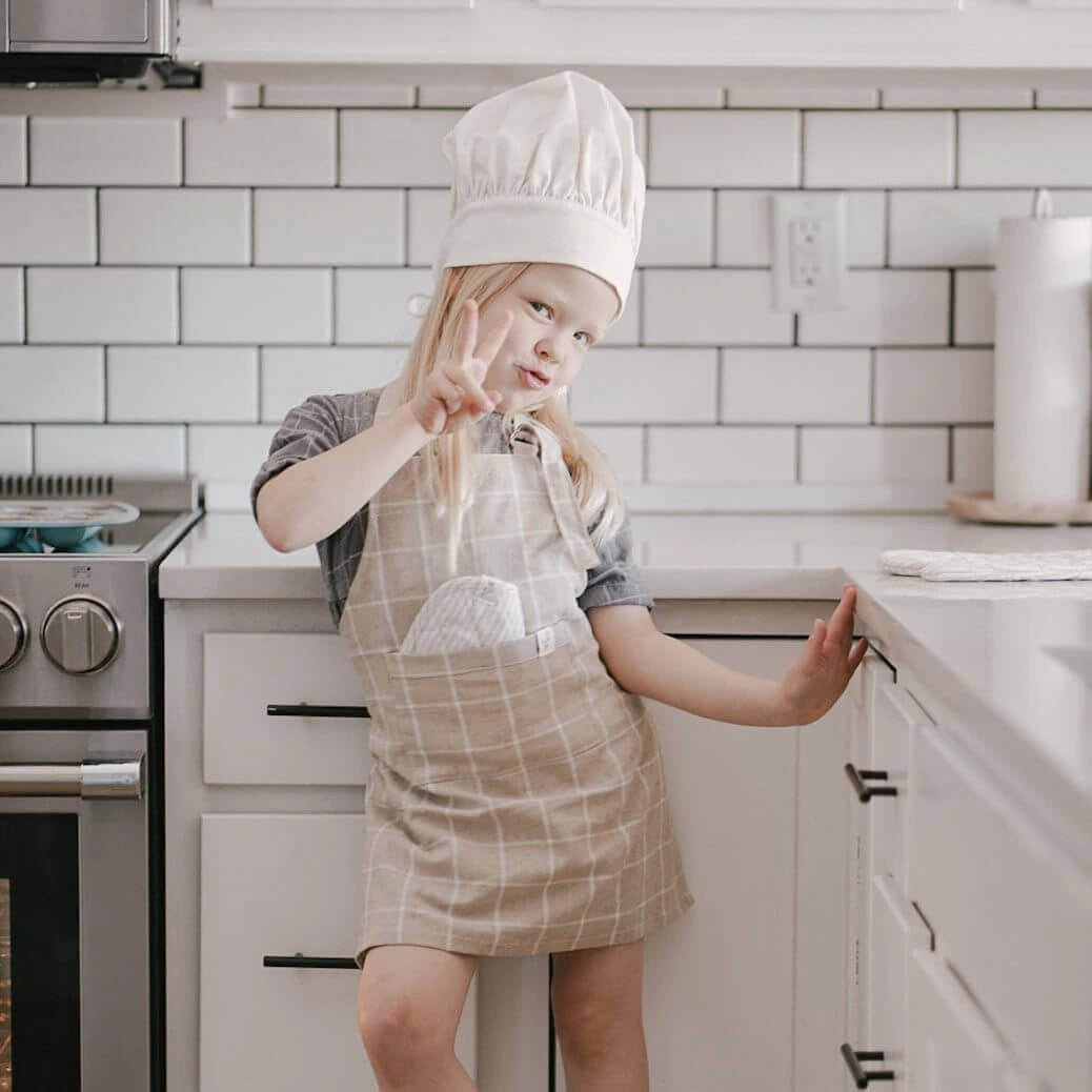 A young girl smiles and makes a peace sign while wearing the Milton & Goose child-sized chef hat, child's apron in tan, and has a play oven mitt stuffed in her apron pocket. 