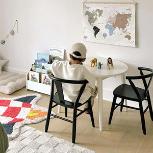 A young boy is shown playing while sitting on a black kid's wood Crescent Chair next to a white Crescent Round Table. 