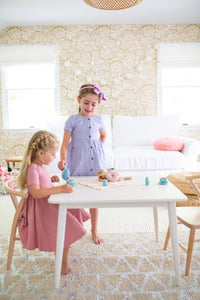 Children play at their Milton & Goose table in white while seated on a child sized crescent chair in natural wood.