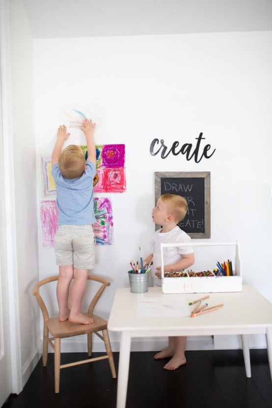 Children hanging art using Milton & Goose Crescent chair in natural with crayons sitting on wooden white child sized table.