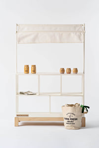 Milton & Goose wooden market stand in white with white canopy. It is styled with Milton & Goose play food and textiles.